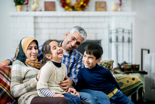 A Muslim mother, father, son and daughter are indoors in a living room. The parents are sitting on the sofa, and the daughter is laughing while being tickled.