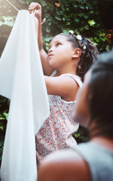 A sweet little girl in yellow rubber rain boots hangs wet clothes on a clothesline in the garden on a hot sunny day