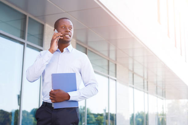 Happy black businessman with smartphone and notepad outdoors. Young african-american salesman working with mobile and papers, standing in urban area cityscape