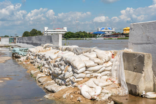 At The Muddy Riverbank,Two Men In Rubber Boots Stand Beside Sacks Filled With Sand Or Gravel,Their Sturdy Legs Ready For The Task At Hand