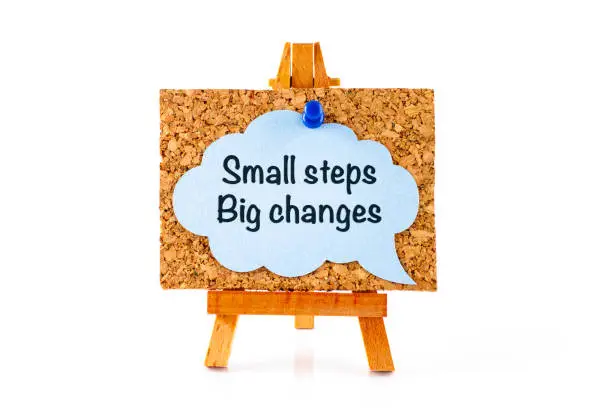 Wooden easel with corkboard and blue speech bubble with phrase Small Steps Big Changes. White background.