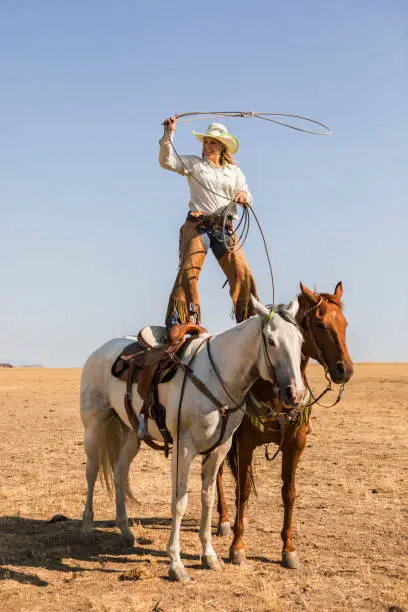 A cowgirl balances on horses as she twirls a lasso.