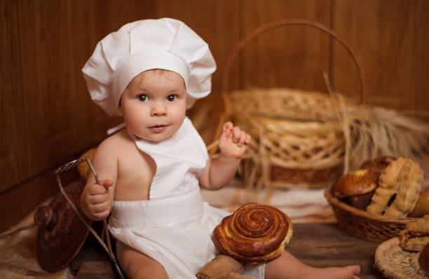 happy little child in the cook costume at the kitchen sitting on the table among baskets with tasty rolls, buns and bread. copy space - ravena imagens e fotografias de stock