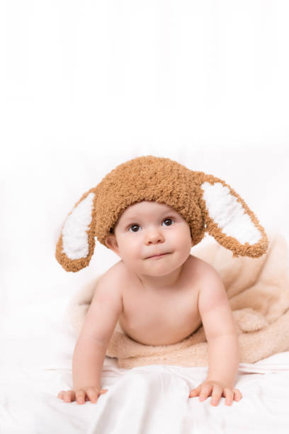 a cute little baby wearing a bunny hat is sitting on the white bed, covered with a towel. white background. screaming child. copy space - ravena imagens e fotografias de stock