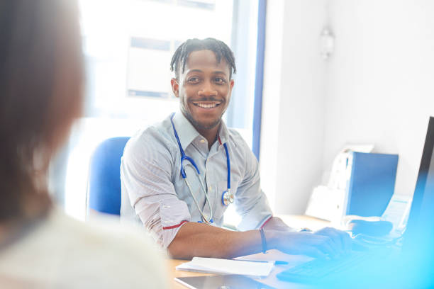 Smiling doctor typing out prescription for patient on computer Junior doctor at his desk in his office in consultation with a patient doctor general practitioner stock pictures, royalty-free photos & images