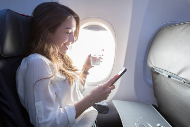 Young woman enjoys in flight beverage and wifi A smiling young woman sits in the window seat of her aircraft.  She holds a beverage in one hand and her smart phone in another as she enjoys the wifi.  Her earbuds rest on a tray in front of her. window seat vehicle stock pictures, royalty-free photos & images