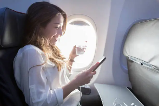 A smiling young woman sits in the window seat of her aircraft.  She holds a beverage in one hand and her smart phone in another as she enjoys the wifi.  Her earbuds rest on a tray in front of her.