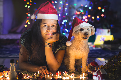 Brazilian woman setting up the Christmas tree, and her dog wearing santa's hat beside several christmas ornaments.
