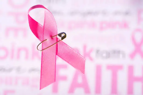 Small pink ribbon with a safety pin, cancer awareness. Background has encouraging words like hope, faith, pink, courage. Copy space towards the right of the ribbon.