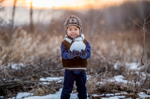 A young Asian boy is standing in a field on a cold winter day. He is wearing a hat, coat and gloves. He is holding a large snowball that he made to build a snowman.