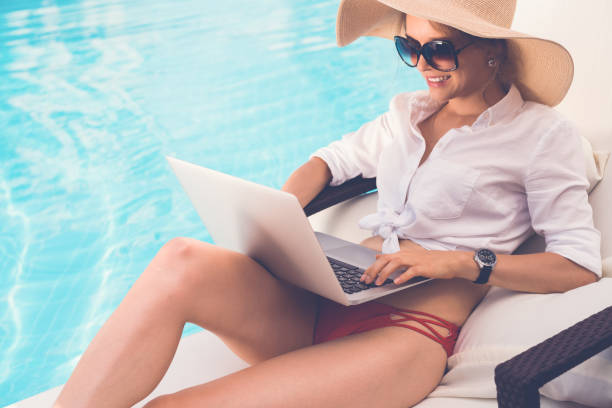 Business ladu Smiling business woman in sunglasses and straw hat sitting by swimming pool and working on laptop chaise longue photos stock pictures, royalty-free photos & images