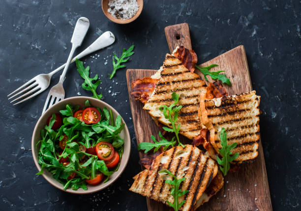 grilled bacon, mozzarella sandwiches on wooden cutting boards and arugula, cherry tomato salad on dark background, top view.delicious breakfast or snack, flat lay - mozzarella tomato sandwich picnic imagens e fotografias de stock