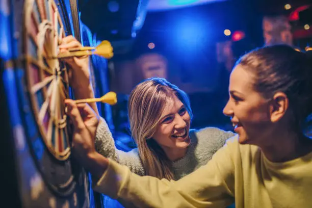 Two female friends having fun while taking darts out of dartboard in a bar.