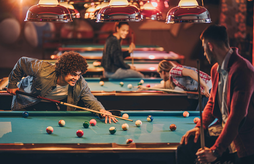 Smiling man aiming at pool ball while playing snooker with friends at entertainment club.