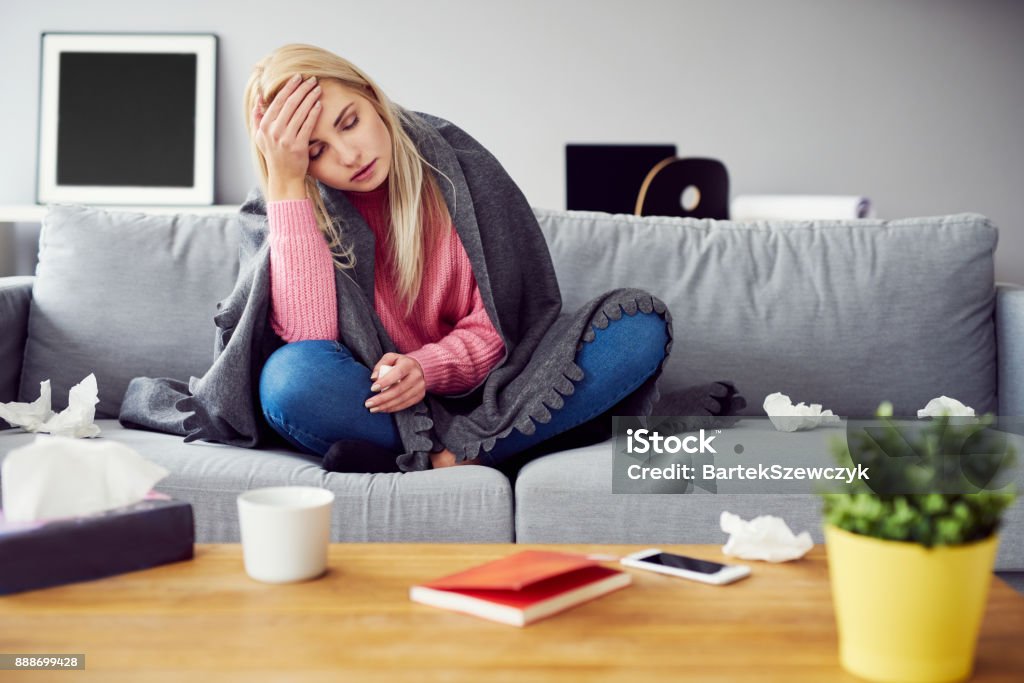 Sick woman with headache sitting under the blanket in living room Cold And Flu Stock Photo