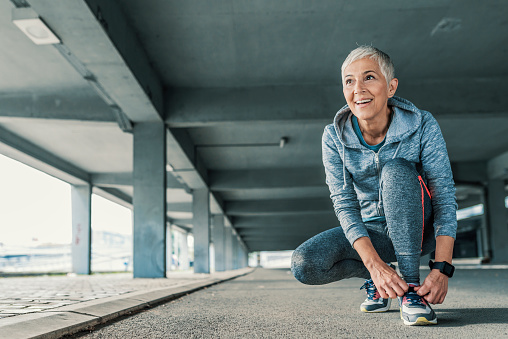 Running shoes - closeup of mature, gray hair woman tying shoe laces and looking forward. Female sport fitness runner getting ready for jogging outdoors  in spring. Senior fitness woman getting ready to start running workout during the sunny day. Fit and sporty short hair woman tying her laces before a run.