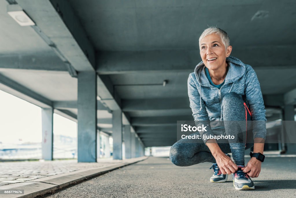 I'm ready to hit the road Running shoes - closeup of mature, gray hair woman tying shoe laces and looking forward. Female sport fitness runner getting ready for jogging outdoors  in spring. Senior fitness woman getting ready to start running workout during the sunny day. Fit and sporty short hair woman tying her laces before a run. Exercising Stock Photo