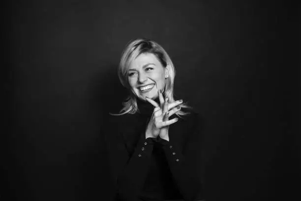 Black and white portrait of a young woman, laughing, looking to the side, holding hands together, against a plain studio background