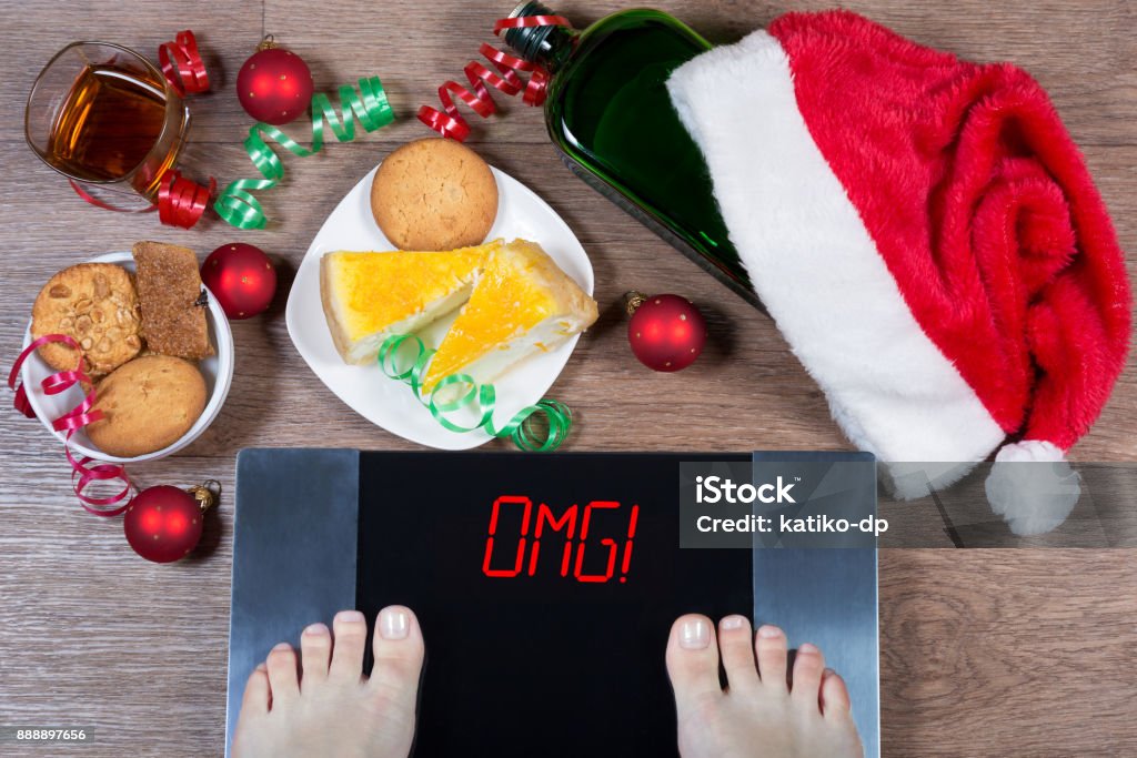 Female feet on digital scales with sign "omg!" surrounded by Christmas decorations, bottle, glass of alcohol and sweets. Consequences of overeating and unhealthy lifestile during holidays. Female feet on digital scales with sign "omg!" surrounded by Christmas decorations, bottle, glass of alcohol and sweets. Consequences of overeating and unhealthy lifestile during holidays. Top view. Holiday - Event Stock Photo