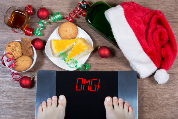 Female feet on digital scales with sign "omg!" surrounded by Christmas decorations, bottle, glass of alcohol and sweets. Consequences of overeating and unhealthy lifestile during holidays. stock photo