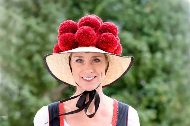 Attractive young woman wearing a traditional Black Forest Bollenhut with its 14 pompoms as she smiles at the camera outdoors against greenery
