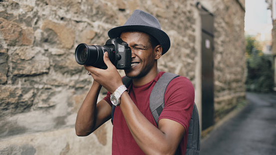 African american happy tourist taking photo on his dslr camera. Young man standing near brick wall of famous building in Europe