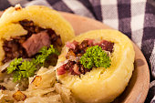 Horizontal photo of potato dumplings filled by smoked pork meat with green parsley twig. Sour cabbage is next to meal on clay plate with checkered towel around. Fried onion is spilled on food.