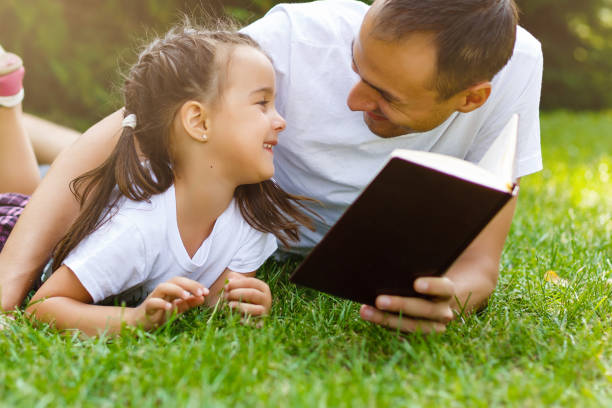 padre e figlio felici che leggono un libro nella natura - outdoors looking at camera little girls child foto e immagini stock