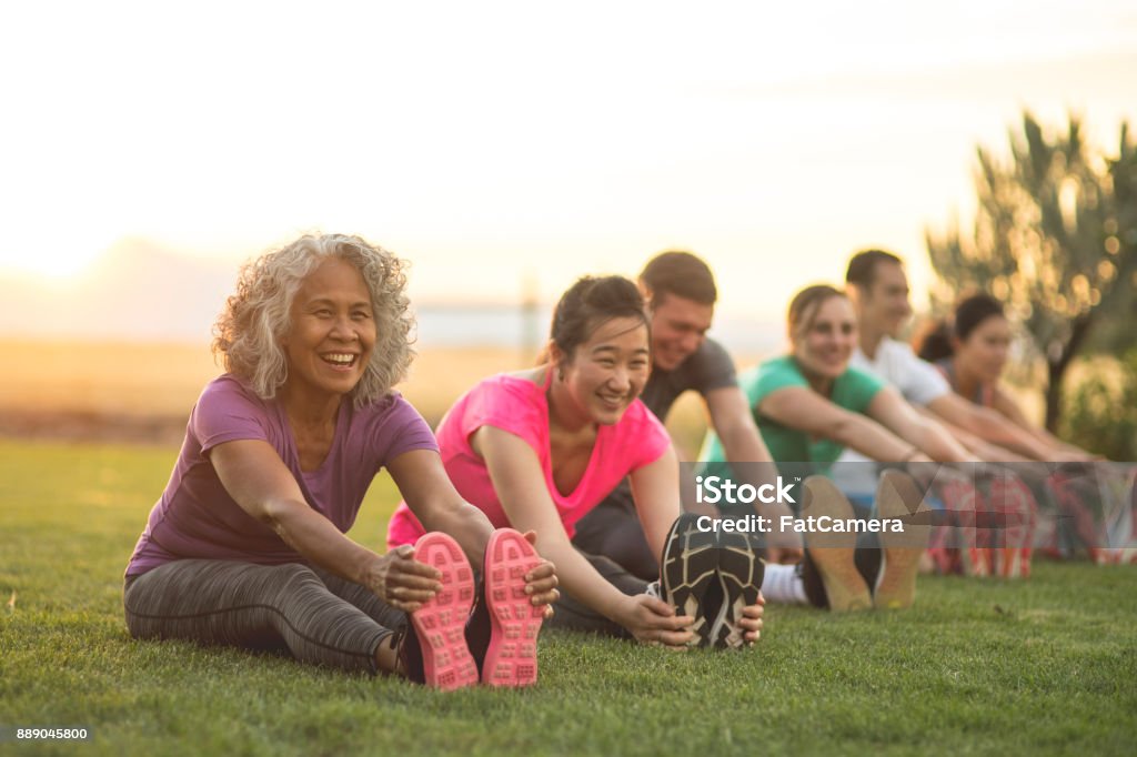 Fitness Class Stretching A group of adults attending a fitness class outdoors are doing leg stretches. The participants are arranged in a line. The focus is on a mature woman who is smiling toward the camera. Exercising Stock Photo