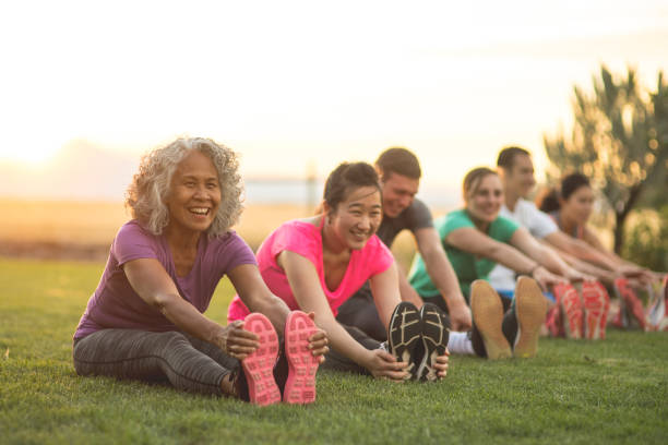 Fitness Class Stretching A group of adults attending a fitness class outdoors are doing leg stretches. The participants are arranged in a line. The focus is on a mature woman who is smiling toward the camera. activity stock pictures, royalty-free photos & images