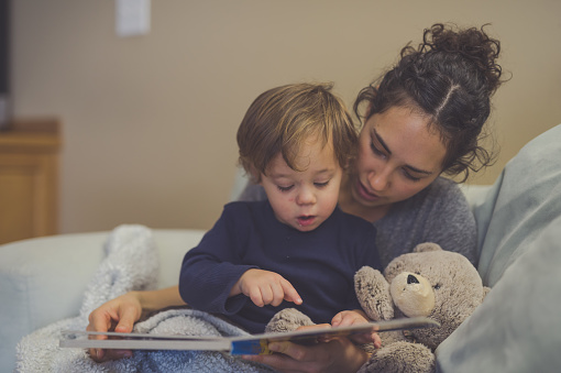 Young Hispanic mom reads a book aloud to her young son, who has food on his face.. They're pointing at the pictures together. A teddy bear is snuggled up with them.
