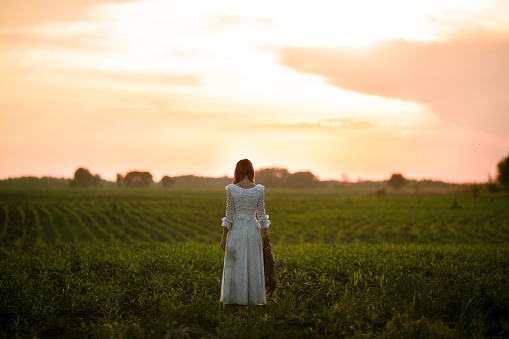 Young woman in long white lace dress on cornfield at sunset. She stands with bouquet of wild flowers in her hands. Back view.