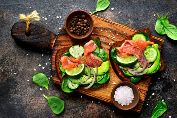 Rye toasts with avocado,cucumber,salmon,and spinach on a wooden cutting board over dark slate,stone or ruasty metal background.Top view with copy space.