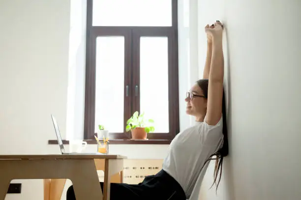 Break at work: female worker stretching hands above head at workplace. Beautiful millennial woman sitting at table in office and relaxing. Simple exercises to relieve stress, office gymnastics concept