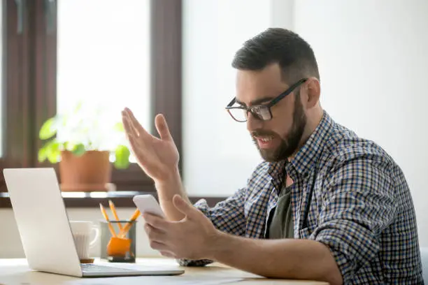 Confused bearded manager in glasses is angry because mobile phone is discharged. Millennial businessman in casual receiving bad news and spam in smartphone