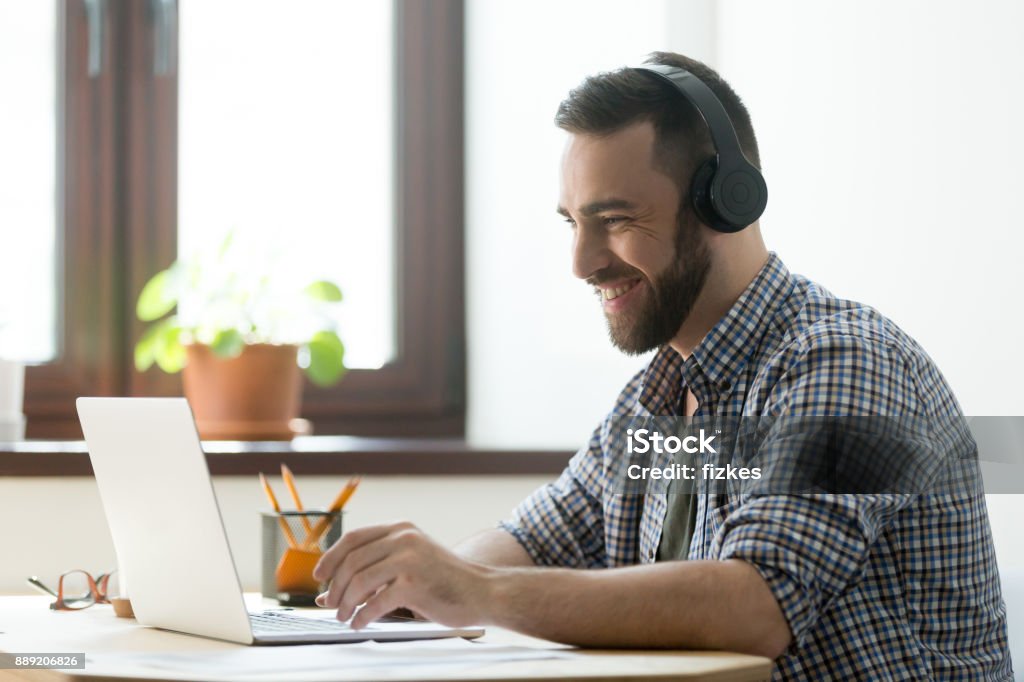 Smiling bearded man in headphones looking on laptop and typing Handsome man with beard listening music in headphones and looking on screen of laptop. Young manager having a rest and playing in computer games or making video call on pc Web Conference Stock Photo