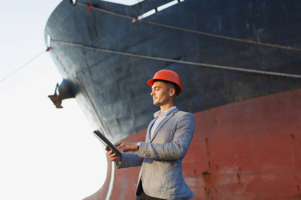 istock Handsome unshaven successful business man in gray suit, protective construction orange helmet holding tablet, standing in sea port against cargo rusty ship background. Male with gadget in sunny day 889390224