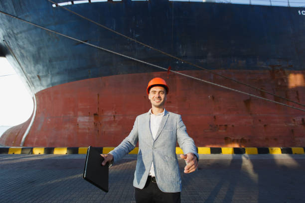 istock Handsome unshaven successful business man in gray suit, protective construction orange helmet holding tablet, standing in sea port against cargo rusty ship on water line background. Male with gadget 889390352
