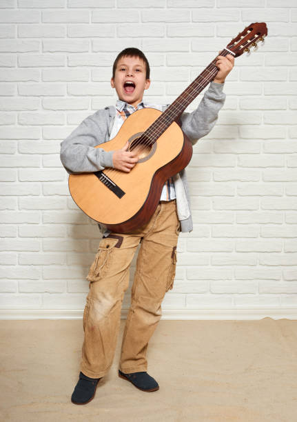 istock boy playing music on guitar, standing at full height, white brick wall background 889491968