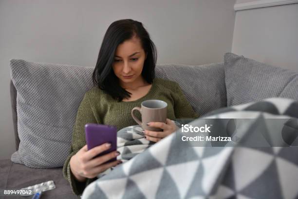 A Sick Young Woman Lies In Bed And Uses A Cell Phone Stock Photo - Download Image Now