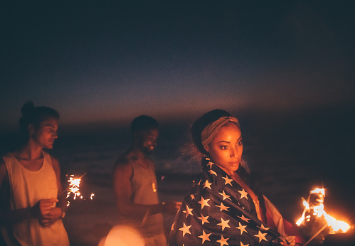 Young multi-ethnic hipster friends with sparklers and American flag at beach celebrating fourth of July