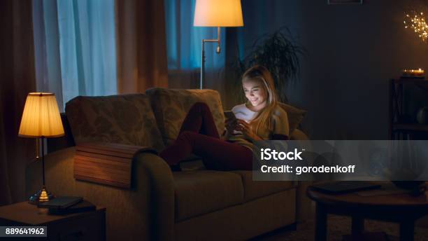 In The Evening Beautiful Young Woman Lies On The Couch And Uses Smartphone Room Looks Warm And Cozy Stock Photo - Download Image Now