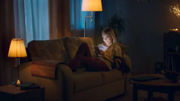 Photo of In the Evening Beautiful Young Woman Lies on the Couch and Uses Smartphone. Room Looks Warm and Cozy.