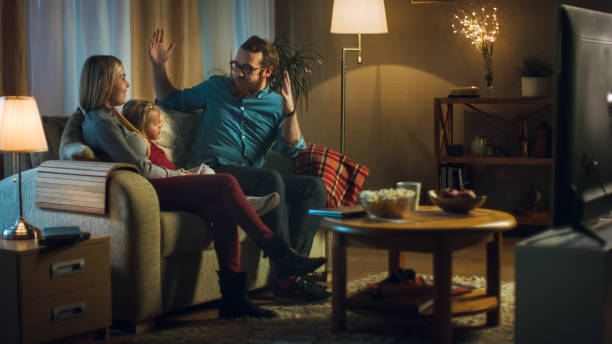 long shot of a father, mother and little girl watching tv. they sit on a sofa in their cozy living room and eat popcorn. it's evening. - apartment television family couple imagens e fotografias de stock