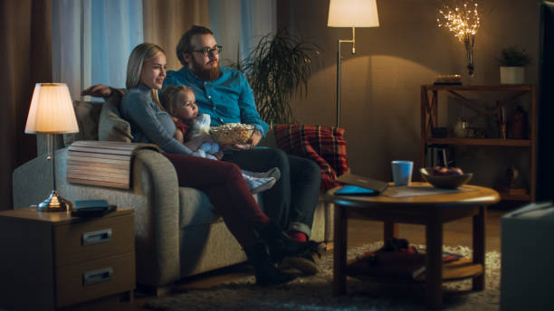 largo tiro de padre, la madre y la niña, viendo la televisión. se sientan en un sofá en su acogedora sala de estar y comen palomitas de maíz. es la noche. - apartment television family couple fotografías e imágenes de stock