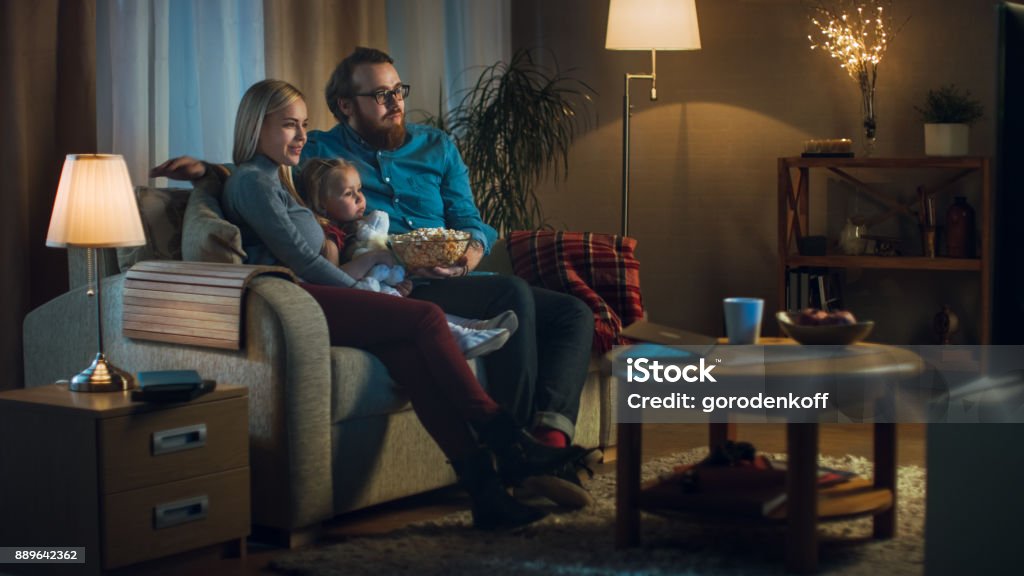 Long Shot of a Father, Mother and Little Girl Watching TV. They Sit on a Sofa in Their Cozy Living Room and Eat Popcorn. It's Evening. Family Stock Photo