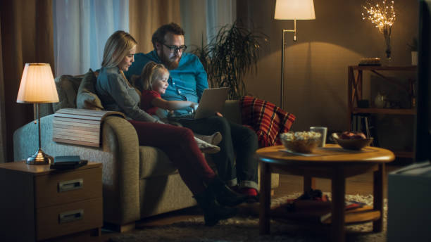 tiro de um pai, mãe e filha assistindo tv. sentam-se em um sofá na sua sala de estar aconchegante, pai detém laptop sobre os joelhos. é noite. - family sofa night indoors - fotografias e filmes do acervo