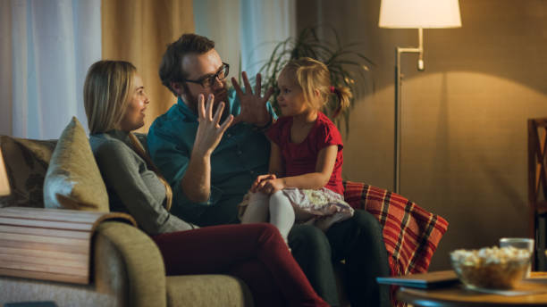 in the evening family spends time together sitting on a couch in a living room. little girl sits on her father's laps and parents tickle her. - apartment television family couple imagens e fotografias de stock
