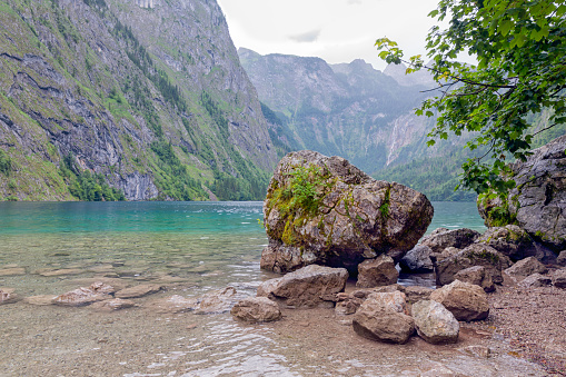 German Konigssee near Berchtesgaden with steep mountain slopes and a big boulder in front lying in clear transparant water