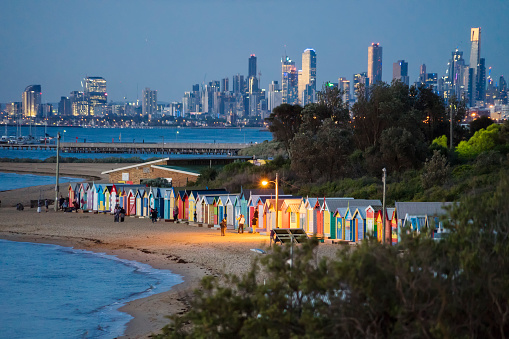 Melbourne, Australia - December 8, 2017: Visitors around beach boxes at Brighton Beach at dusk. Skyscrapers in the city visible in the distance.